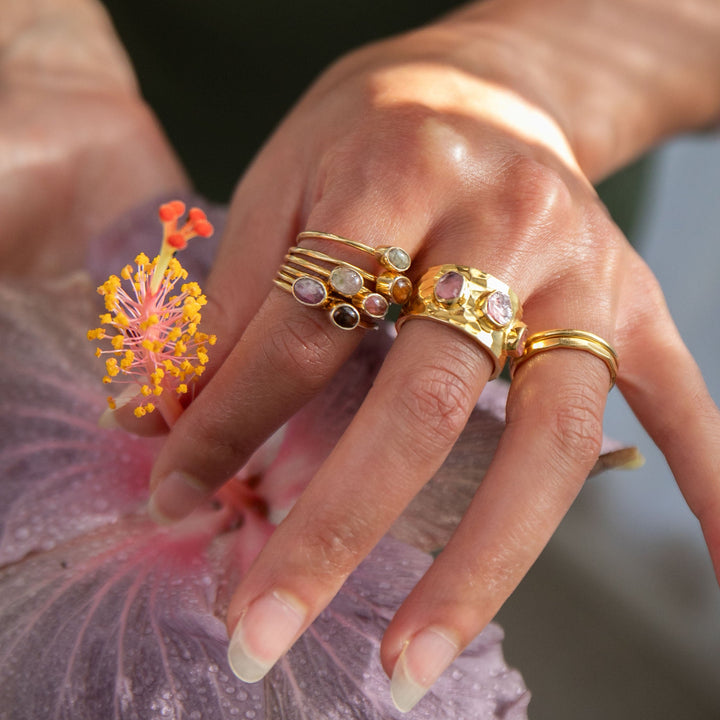 A close-up of a person's hand wearing multiple gold rings, resting on a pink flower. The rings feature gold bands with various stone and crystal accents.
