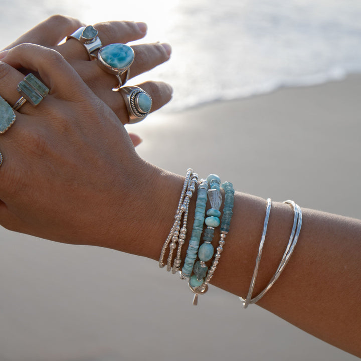 A person's wrist wearing multiple bracelets with Aquamarine, Larimar, and Peruvian Opal, alongside Hill Tribe Silver beads, against a beach backdrop.